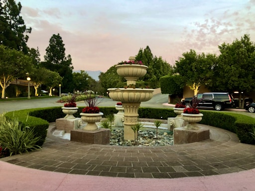 A beautifully manicured garden setting features a prominent stone fountain surrounded by brick paving and circular hedges. The fountain is adorned with potted red flowers, and a driveway in the background is lined with trees and bushes under a soft pink and blue evening sky.