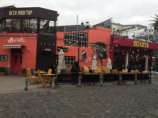 Outdoor seating area on a cobblestone street featuring a vibrant mural of a woman with a bird on an orange-pink wall. Patio furniture includes wooden tables and chairs with umbrellas. The establishment signs read 'BEER ROOFTOP,' 'Brewery,' and 'CREPAS,' accompanied by string lights and decorative plantings.