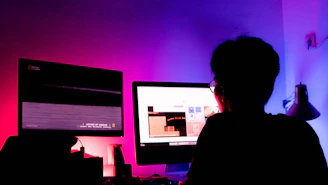 A sleek dark-themed workspace with pink and cyan lighting, showing a game producer reviewing project plans on multiple screens.