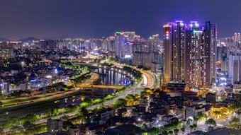 A vibrant cityscape at night, featuring tall skyscrapers illuminated by colorful lights. A river runs through the city, reflecting the bright lights from the buildings and street lamps. The scene is lively with bustling streets, and various buildings in the foreground leading up to the high-rise structures.