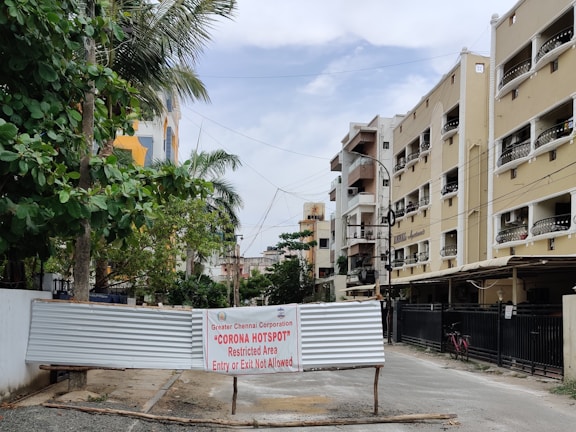 A residential street scene features multi-story apartments lining both sides of a road, with lush green trees on the left. A metal barricade is placed across the road with a sign from the Greater Chennai Corporation indicating a 'Corona Hotspot,' marked as a restricted area with no entry or exit allowed.