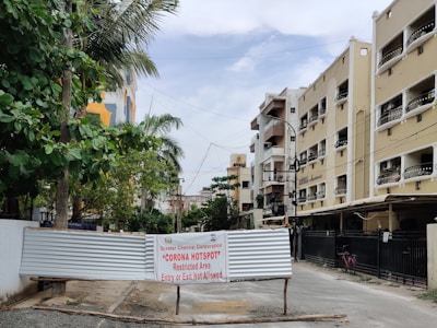 A residential street scene features multi-story apartments lining both sides of a road, with lush green trees on the left. A metal barricade is placed across the road with a sign from the Greater Chennai Corporation indicating a 'Corona Hotspot,' marked as a restricted area with no entry or exit allowed.