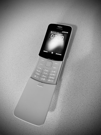A black and white image of a nostalgic flip phone placed on a textured surface. The phone is open, revealing a keypad with numeric and function keys. The screen shows a digital clock display.