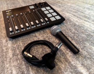 A sound mixing console is placed on a carpeted surface, featuring multiple sliders and buttons. Accompanying the console is a handheld microphone and a coiled audio cable nearby.