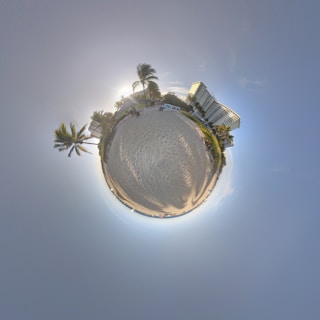 A spherical panoramic view of a sandy beach with palm trees and a building in the background. The sky is clear, and the sun is setting, casting long shadows.