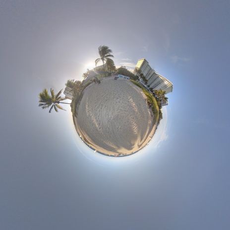 A spherical panoramic view of a sandy beach with palm trees and a building in the background. The sky is clear, and the sun is setting, casting long shadows.