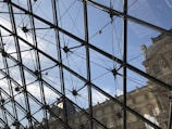 Glass and metal structure with a geometric pattern giving a view of a clear blue sky and part of a classical building facade with detailed stonework.
