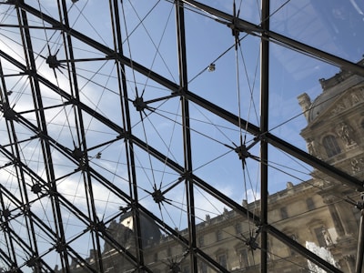 Glass and metal structure with a geometric pattern giving a view of a clear blue sky and part of a classical building facade with detailed stonework.