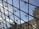 Glass and metal structure with a geometric pattern giving a view of a clear blue sky and part of a classical building facade with detailed stonework.