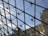 Glass and metal structure with a geometric pattern giving a view of a clear blue sky and part of a classical building facade with detailed stonework.