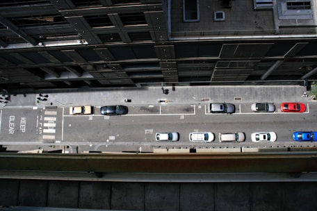 An aerial view of a city street lined with parked cars on one side. Several vehicles including a red car and a blue car are parked in marked spaces. Pedestrians are visible on the sidewalk, and road markings such as a pedestrian crossing and 'KEEP CLEAR' text are present.
