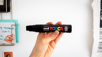 A hand is holding a large black marker with colorful branding that says 'uni Posca'. The background includes various papers on a wall, one of which has handwritten notes. The focus is on the marker with sharp details of the hand and the product's label.