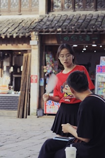 A young woman in a red shirt with a cartoon character is standing and holding a fan, talking to a seated man dressed in black with a white cup in front of him. They are in an outdoor setting with traditional architecture in the background, including a tiled roof and wooden details, suggesting a market or historic area.