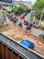 A busy street scene with multiple tuk-tuks and motorcycles moving along the road. People are riding scooters, and a person wears traditional attire, carrying a red bag. The environment is urban with visible wires overhead, trees and buildings in the background, including a visible blue car parked roadside.