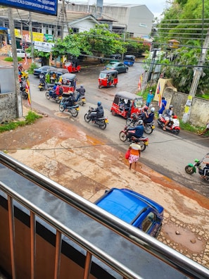 A busy street scene with multiple tuk-tuks and motorcycles moving along the road. People are riding scooters, and a person wears traditional attire, carrying a red bag. The environment is urban with visible wires overhead, trees and buildings in the background, including a visible blue car parked roadside.