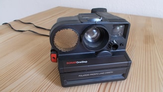 An old-fashioned Polaroid camera with the label SONAR OneStep is resting on a light wooden surface. The camera features a prominent circular disk on the left, possibly a sensor or flash, and a large lens in the center. The camera has a classic black design, with some red and gold accents. Its cord is visible in the background, laying on the wooden table.