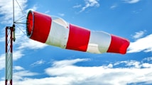 A red and white windsock extends horizontally, indicating wind direction and speed. It is attached to a metal pole structure against a backdrop of a bright blue sky with scattered clouds.