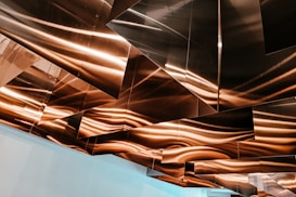 Geometric metal panels are installed on a ceiling, reflecting warm light and creating an abstract pattern with wavy lines.