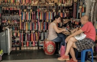 A small shop filled with colorful tassels and accessories, where a woman is engaged in conversation with an older man. The woman is sitting on a decorative drum, while the man occupies a blue plastic stool. The shelves behind them are packed with various trinkets and rolls of thread in vibrant hues.