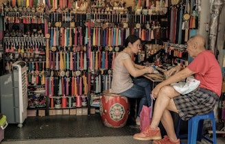 A small shop filled with colorful tassels and accessories, where a woman is engaged in conversation with an older man. The woman is sitting on a decorative drum, while the man occupies a blue plastic stool. The shelves behind them are packed with various trinkets and rolls of thread in vibrant hues.