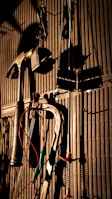 Various tools including a hammer, wrenches, and cables hang on a vertically striped surface. The light casts strong shadows, creating a dramatic effect with the tools silhouetted against the background.