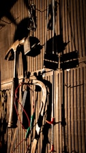 Various tools including a hammer, wrenches, and cables hang on a vertically striped surface. The light casts strong shadows, creating a dramatic effect with the tools silhouetted against the background.