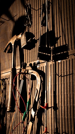 Various tools including a hammer, wrenches, and cables hang on a vertically striped surface. The light casts strong shadows, creating a dramatic effect with the tools silhouetted against the background.