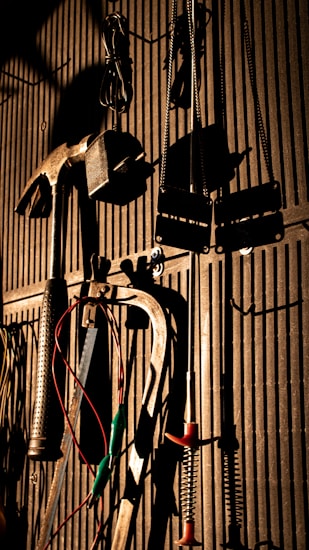 Various tools including a hammer, wrenches, and cables hang on a vertically striped surface. The light casts strong shadows, creating a dramatic effect with the tools silhouetted against the background.
