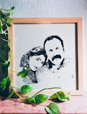 A framed line-art portrait of two people, a man and a woman, is leaning against a textured wall on a marble surface. Green vine leaves from an indoor plant drape over and around the frame, adding a natural element to the scene.