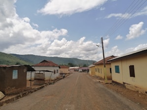 A rural road lined with a few single-story buildings on both sides, some with corrugated metal roofs. The road extends into the distance, with utility poles and power lines on one side. The sky is partly cloudy with lush green hills visible in the background.
