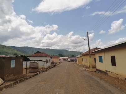 A rural road lined with a few single-story buildings on both sides, some with corrugated metal roofs. The road extends into the distance, with utility poles and power lines on one side. The sky is partly cloudy with lush green hills visible in the background.
