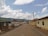 A rural road lined with a few single-story buildings on both sides, some with corrugated metal roofs. The road extends into the distance, with utility poles and power lines on one side. The sky is partly cloudy with lush green hills visible in the background.
