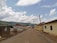 A rural road lined with a few single-story buildings on both sides, some with corrugated metal roofs. The road extends into the distance, with utility poles and power lines on one side. The sky is partly cloudy with lush green hills visible in the background.