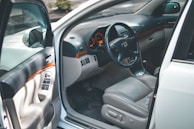 The focus is on the interior of a car with an open driver's side door, revealing a clean and well-maintained beige leather seat. The dashboard includes a steering wheel with a car logo, analog speedometer and gauges, and polished wood trim accents. Controls for windows and mirrors are visible on the door panel.