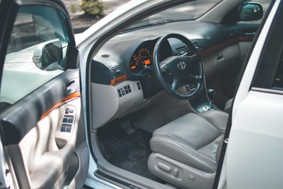 The focus is on the interior of a car with an open driver's side door, revealing a clean and well-maintained beige leather seat. The dashboard includes a steering wheel with a car logo, analog speedometer and gauges, and polished wood trim accents. Controls for windows and mirrors are visible on the door panel.