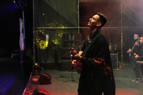 A musician holds a guitar on stage, gazing upward as if deep in thought. He is draped in a poncho-like garment with colorful patterns. Behind him, other band members and sound equipment are visible. The stage is dimly lit with spotlights casting dramatic shadows.