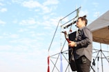 A man wearing a microphone headset is playing a guitar on an outdoor stage. He is standing near scaffolding and a tent, with blue sky and light clouds in the background. The man appears to be engrossed in his performance.
