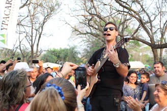 A musician wearing sunglasses performs energetically with an acoustic guitar while surrounded by an enthusiastic crowd in an outdoor setting. People are capturing the moment with their phones, and trees with sparse leaves are visible in the background along with some white tents.