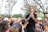 A musician wearing sunglasses performs energetically with an acoustic guitar while surrounded by an enthusiastic crowd in an outdoor setting. People are capturing the moment with their phones, and trees with sparse leaves are visible in the background along with some white tents.