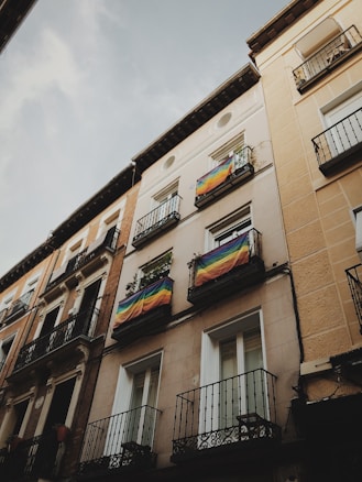 A tall urban building with multiple balconies, each adorned with vivid rainbow flags. The facade is composed of beige and brown hues, with intricate metal railings featuring classic designs. The scene gives a sense of cultural vibrancy within a quiet street.