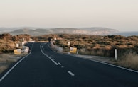 A winding road stretches into the distance through a hilly, coastal landscape. The road is flanked by low vegetation and occasional road signage, including speed limit signs indicating 60 km/h. The sky is clear, and the background features a rugged coastline with cliffs meeting the sea.