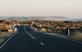 A winding road stretches into the distance through a hilly, coastal landscape. The road is flanked by low vegetation and occasional road signage, including speed limit signs indicating 60 km/h. The sky is clear, and the background features a rugged coastline with cliffs meeting the sea.