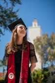A graduate stands outdoors, smiling and wearing a black cap and gown with a red stole. The background shows a blue sky with some trees and a building with a tower.