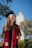 A graduate stands outdoors, smiling and wearing a black cap and gown with a red stole. The background shows a blue sky with some trees and a building with a tower.