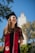 A graduate stands outdoors, smiling and wearing a black cap and gown with a red stole. The background shows a blue sky with some trees and a building with a tower.