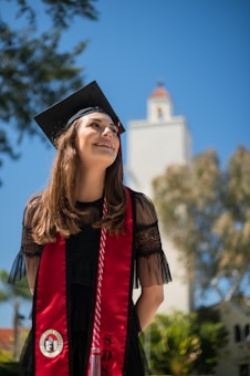 A graduate stands outdoors, smiling and wearing a black cap and gown with a red stole. The background shows a blue sky with some trees and a building with a tower.
