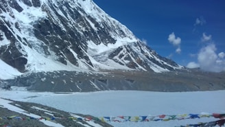 A majestic snowy mountain range under a bright blue sky with scattered clouds. In the foreground, colorful prayer flags are strung across, and a frozen body of water is visible at the base of the mountains.