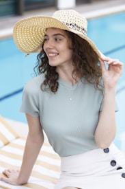 A young woman with curly hair wears a wide-brimmed straw hat while sitting by a swimming pool. She is smiling and looking to the side, dressed in a light-colored, ribbed shirt and a skirt with button details. The setting appears to be relaxed and summery, with bright, natural lighting.