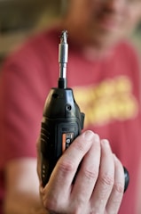 A person is holding a black cordless drill with a metal drill bit attached, prominently displayed in focus. The background is blurred, including a person wearing a red shirt.