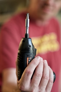 A person is holding a black cordless drill with a metal drill bit attached, prominently displayed in focus. The background is blurred, including a person wearing a red shirt.